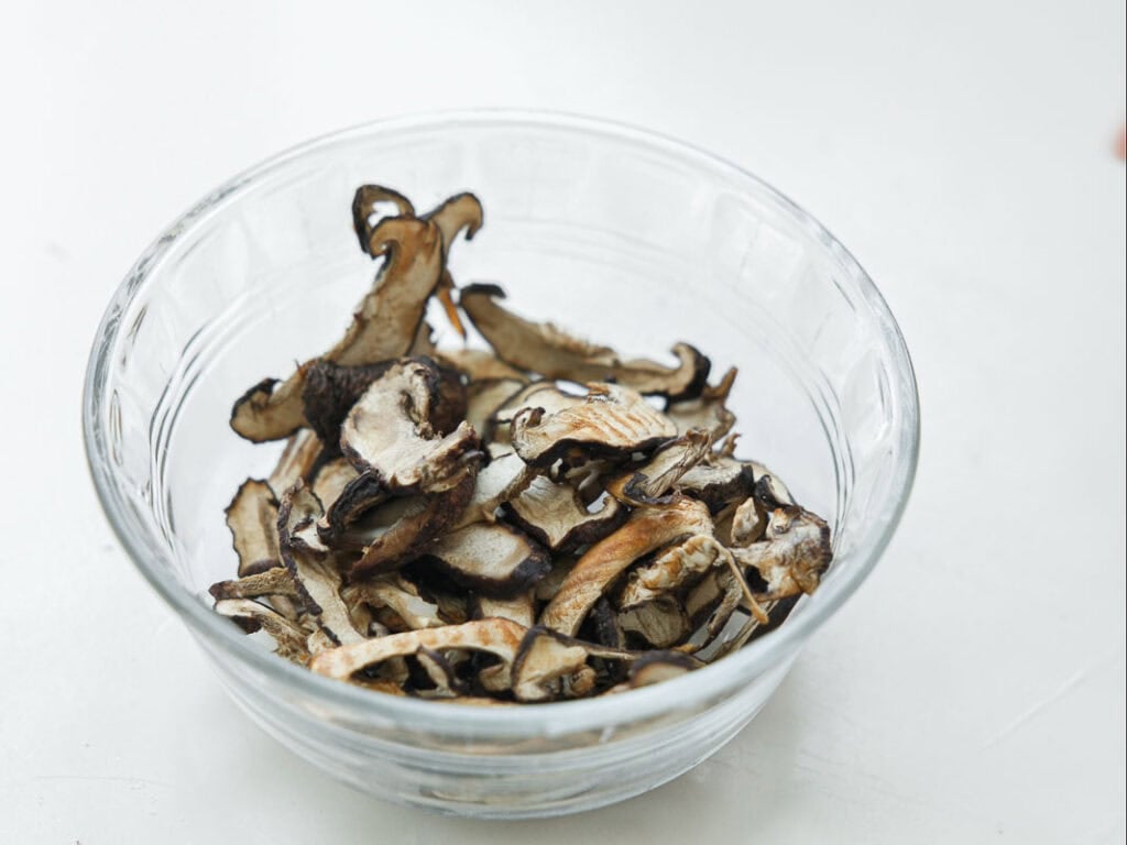 A clear glass bowl filled with dried mushroom slices sits on a white surface.